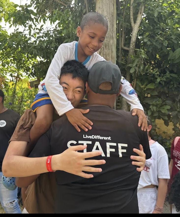 A smiling child sits on a volunteer's shoulders while a LiveDifferent staff member is hugged by the team